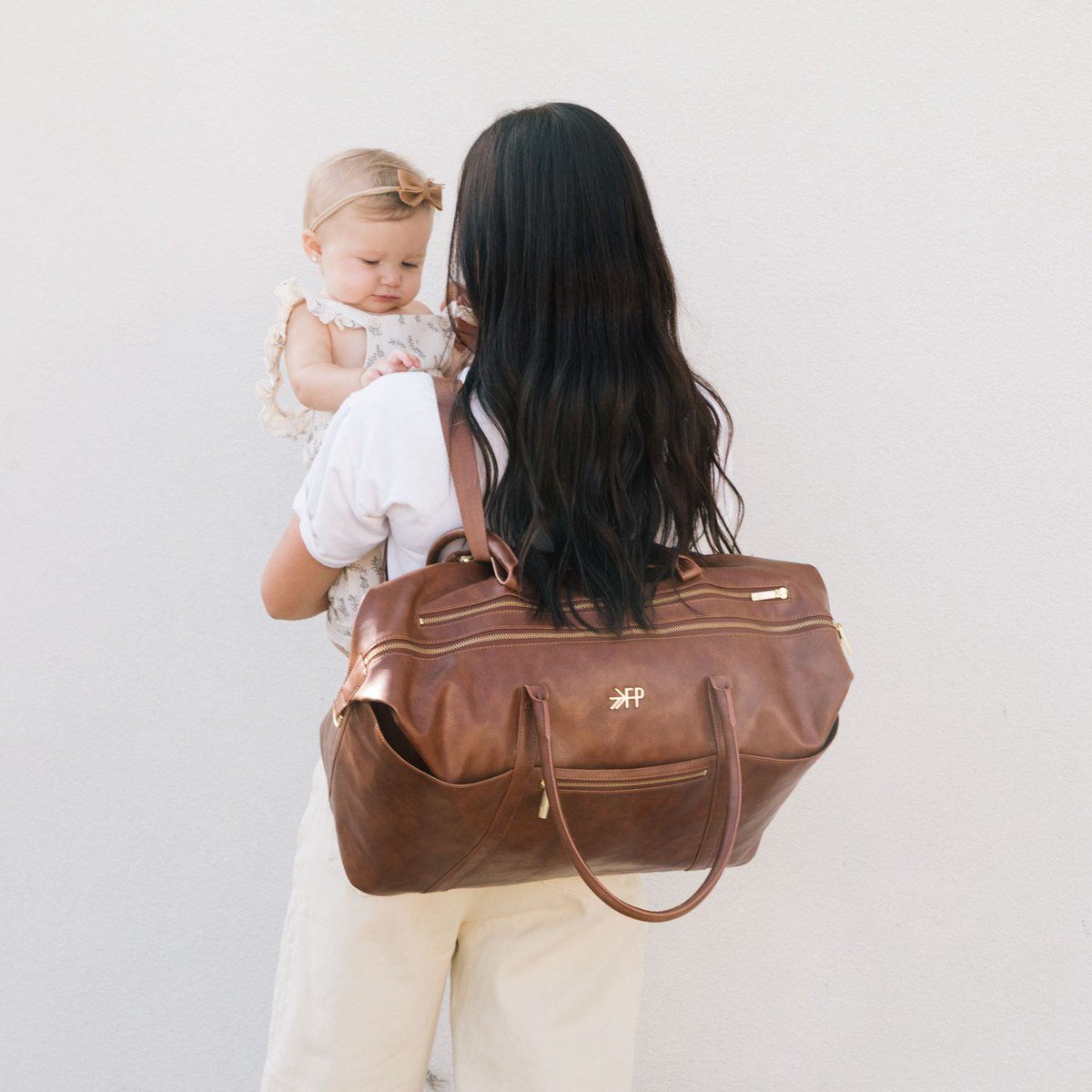 Woman holding a baby and a brown leather bag with a visible brand logo on a plain background
