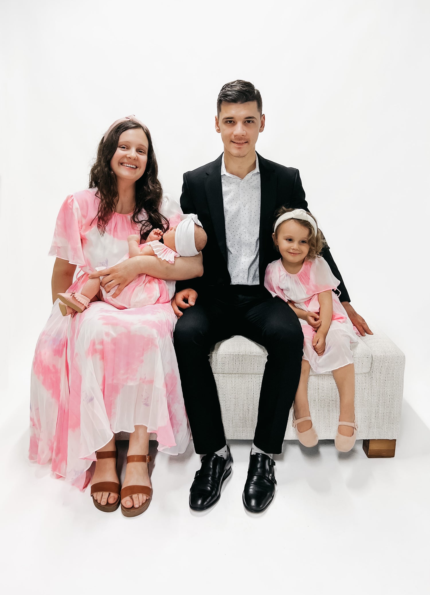 Ruth, founder of Little Mae Co sitting on a stool and holding her baby. Her husband is sitting beside her wearing a black suit and their daughter sits beside wearing a pink dress and headband.